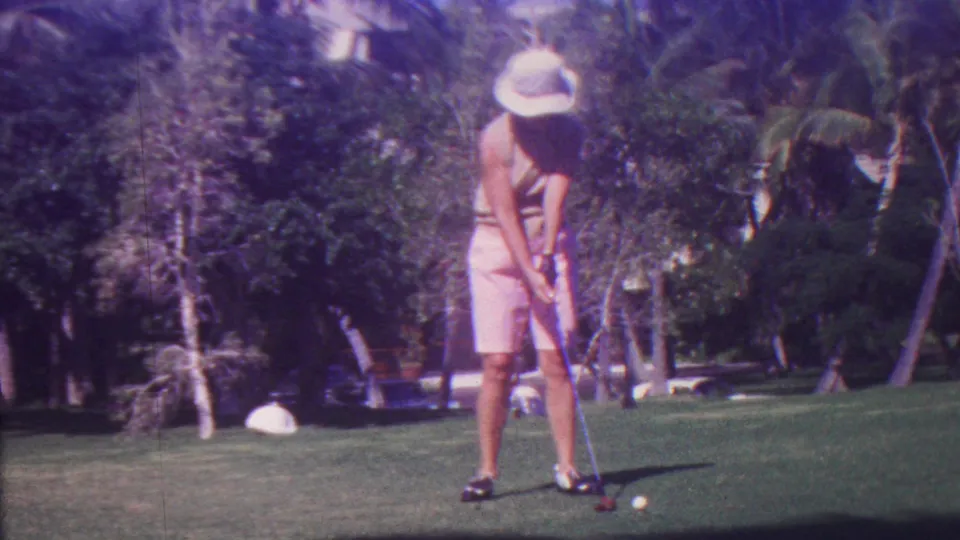 In 1962 Hawaii, the golfer bends slightly to address a white ball on the fairway, framed by tropical trees and a sunlit green.