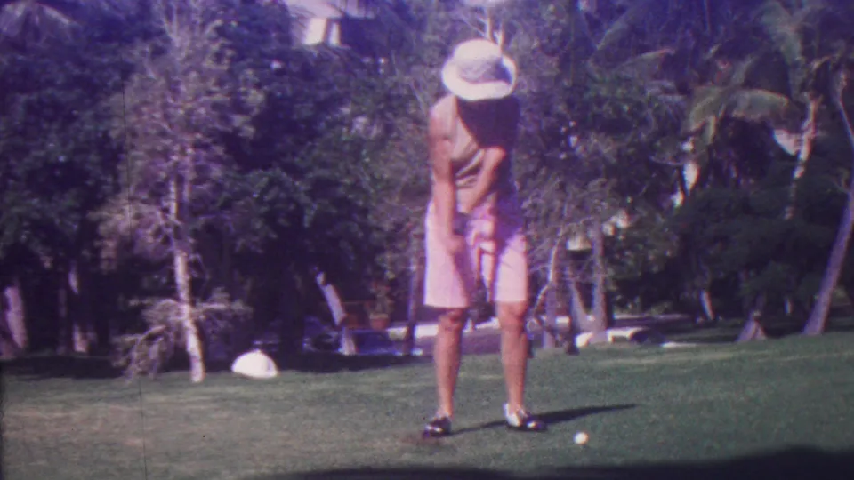 In 1962 Hawaii, the golfer stands over the ball with a club in hand on a green course, with palms and dense vegetation in the background.