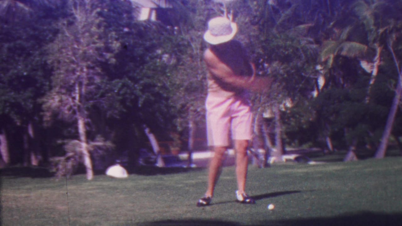 In metadata-supported 1962 Hawaii, a golfer in a wide-brimmed hat and pink shorts stands on a sunlit tropical golf course with a club raised before a swing.
