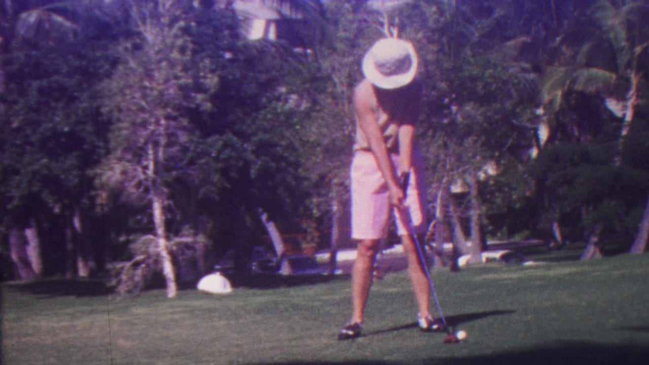 In the Hawaii golf-course setting, the golfer bends at address with a club near a white ball, framed by palms, dense greenery, and bright sunlight.