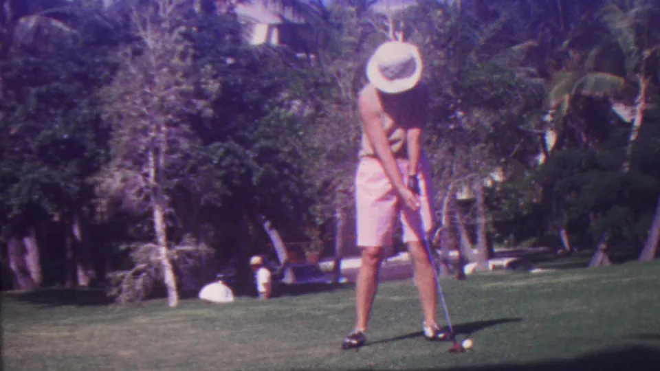 The golfer appears mid-follow-through on a lush Hawaii golf course, with pink shorts, a wide hat, a visible club, and tropical plantings behind the fairway.