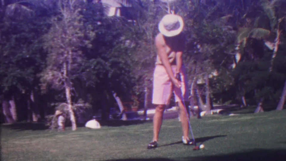 A golfer in a light hat and pink shorts stands over a white ball on a sunlit Hawaii golf course, with palms and dense greenery forming the background.