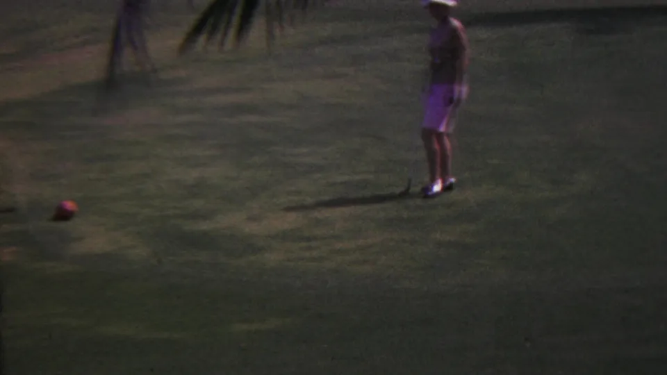 1962 Hawaii 8mm frame showing a woman on a tropical golf green preparing near a ball with palm trees nearby.