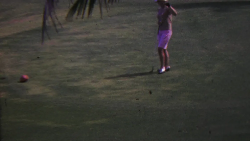 1962 Hawaii 8mm frame showing a woman in light clothing holding a golf club on a palm-lined green with a ball visible nearby.