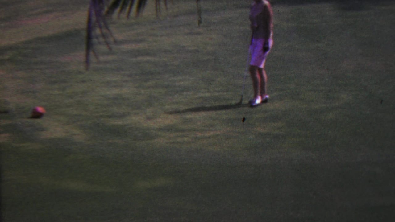 In 1962 Hawaii, a woman in a light outfit stands on a tropical golf green with a red golf ball on the grass and palm fronds hanging above.