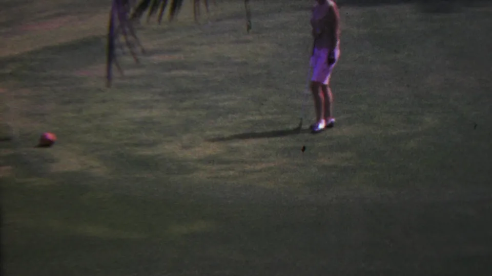 In 1962 Hawaii, a woman in a light outfit stands on a tropical golf green with a red golf ball on the grass and palm fronds hanging above.