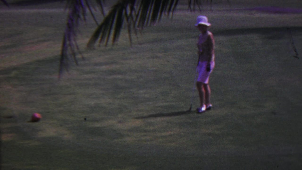 In 1962 Hawaii, a woman wearing a white sun hat stands on a golf course near a red ball, with palm fronds crossing the upper frame.