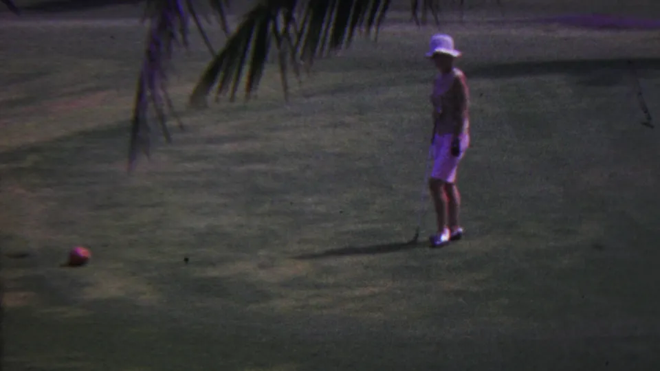 In 1962 Hawaii, a woman wearing a white sun hat stands on a golf course near a red ball, with palm fronds crossing the upper frame.