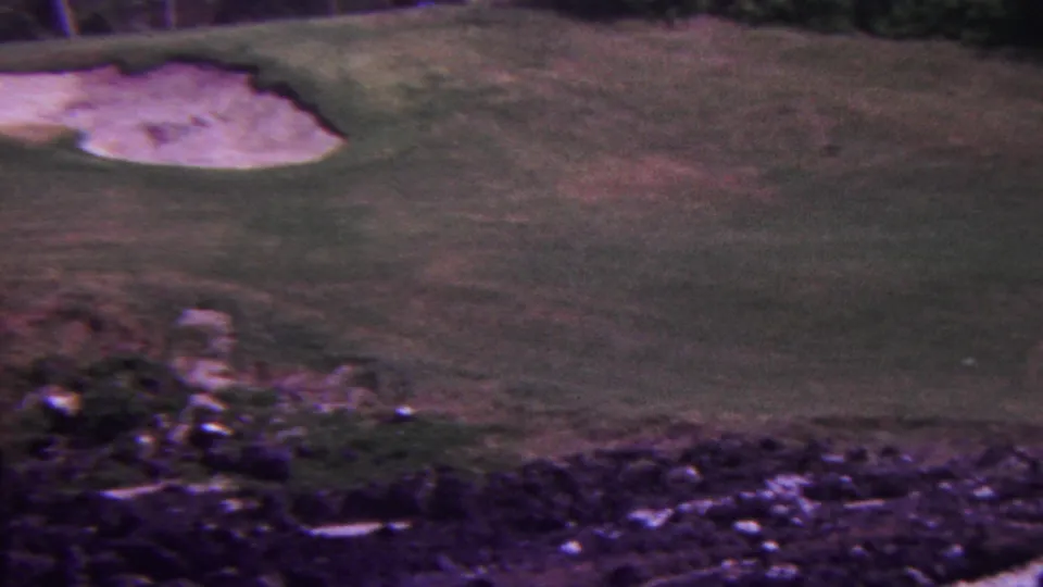 1962 Hawaii 8mm frame showing a golf course fairway with pale sand bunkers, rocky terrain in the foreground, and dark vegetation along the upper edge.