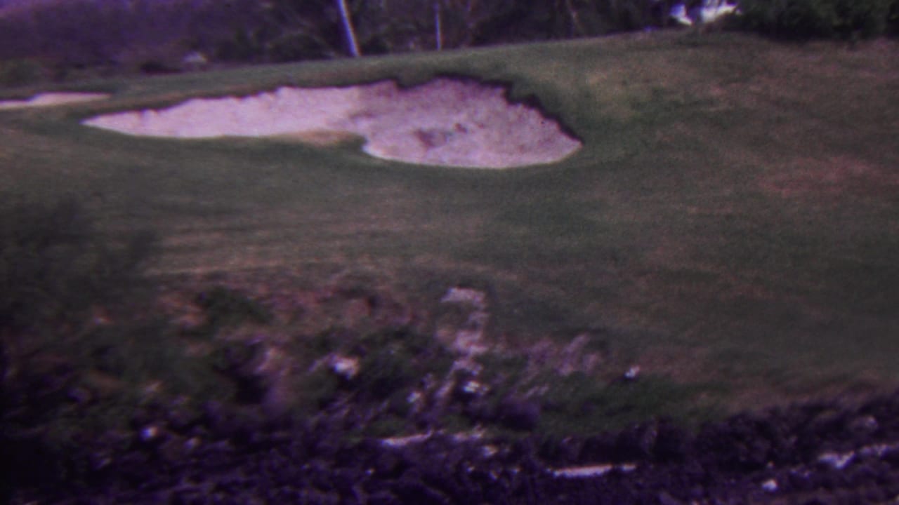 1962 Hawaii 8mm frame of a golf course green with two pale bunkers and a distant golfer or course visitor in light-colored clothing near the right side.