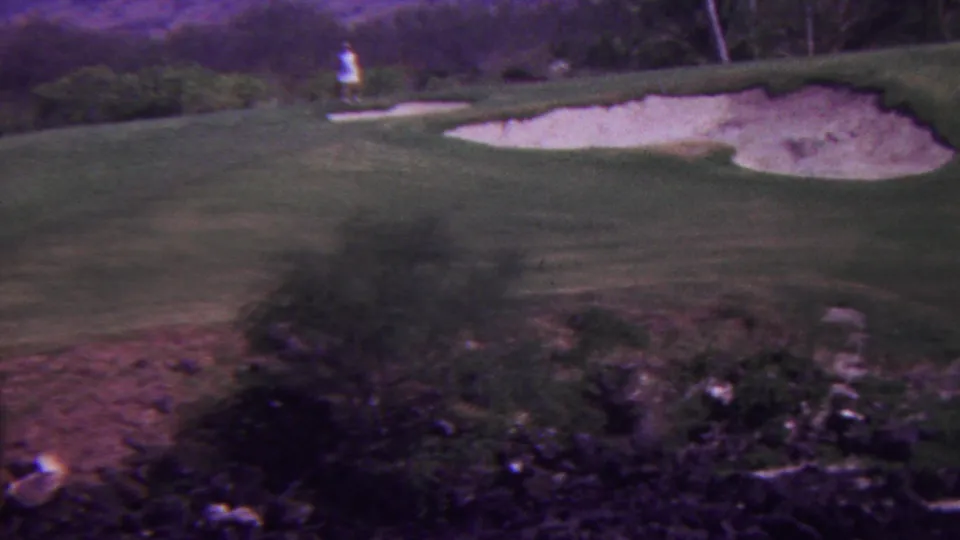 1962 Hawaii 8mm frame showing the course from an elevated angle, with pale bunkers, shrubs and rocks in the foreground, and a distant standing figure on the green.