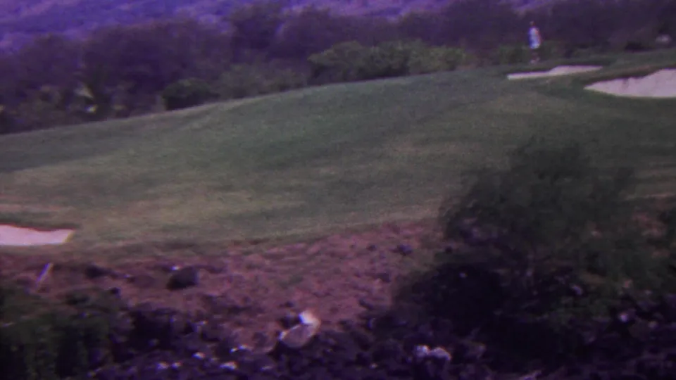 1962 Hawaii 8mm frame with a wide view of a green fairway, a small sand trap, rocky reddish foreground terrain, shrubs, and a distant person in light clothing.
