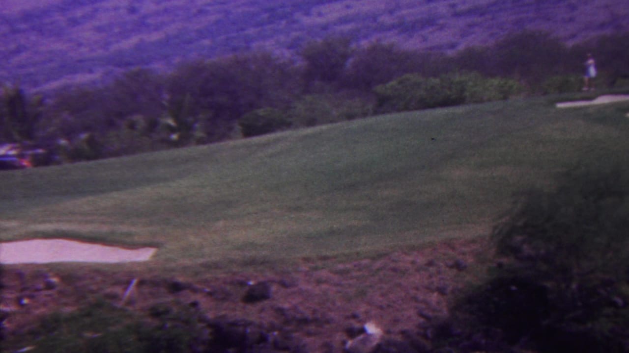 1962 Hawaii 8mm frame showing a green golf course with a white sand trap, palm trees, distant hills, and a small golfer-like figure near the upper right.