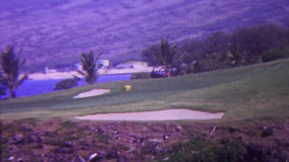 1962 Hawaii 8mm frame showing a landscaped golf course with palms, a bright blue water feature, sand traps, and distant hillside terrain.