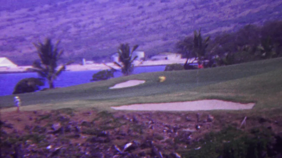 1962 Hawaii 8mm frame showing a wide view of a tropical golf course with multiple sand traps, palms, a blue water feature, rocky foreground, and hills behind.