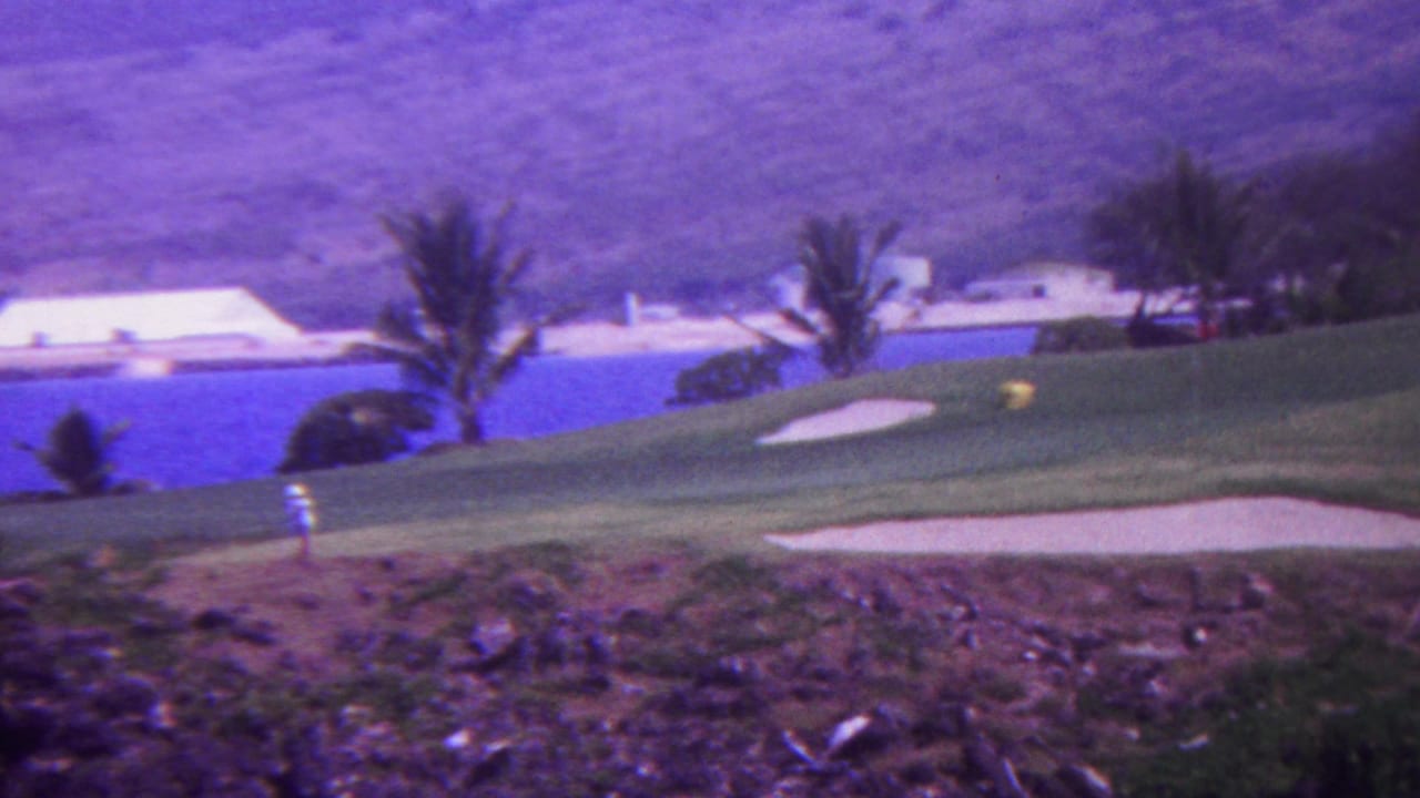 1962 Hawaii home-movie frame showing an ocean-view golf course with palms, sand bunkers, rocky foreground terrain, and a distant white waterfront building.