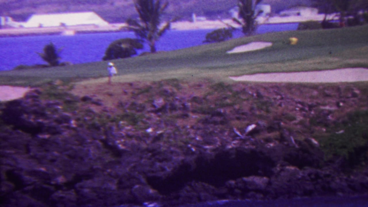 1962 Hawaii 8mm frame with a distant golfer walking across an ocean-view green bordered by sand bunkers, palms, and rocky terrain.