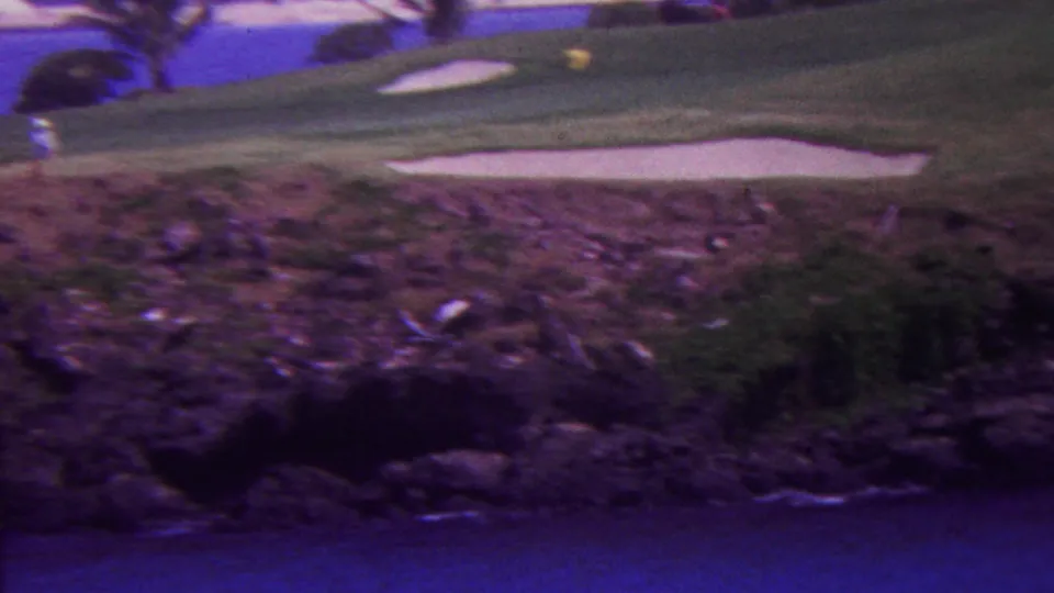 1962 Hawaii home-movie frame showing sand bunkers on a sloping golf green, a small distant golfer, palms, rocky shoreline terrain, and water at the bottom edge.