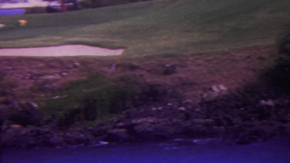 1962 Hawaii 8mm frame showing pale sand bunkers and green turf above dark rocky foreground, with blue water along the lower edge and a small course marker visible.