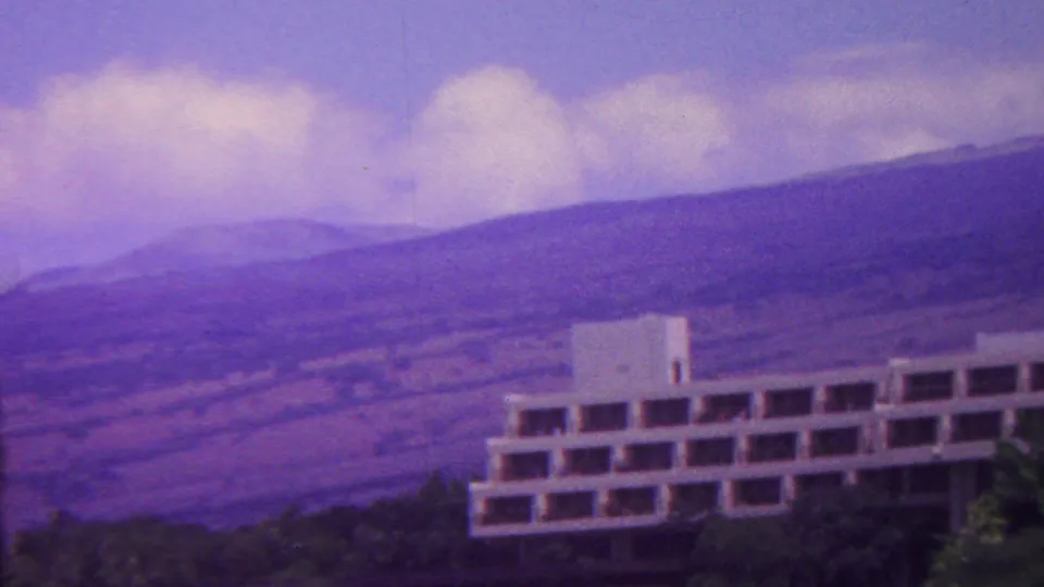 1962 Hawaii 8mm frame showing a white multi-story building set against broad mountain slopes under a cloudy sky.