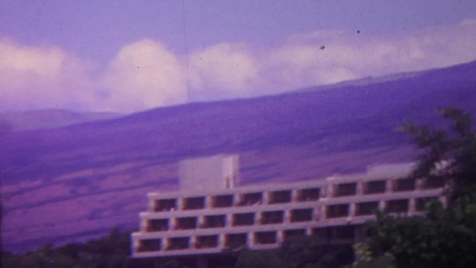1962 Hawaii home-movie frame with a white low-rise multi-story building below purple-toned mountains and a cloudy sky.