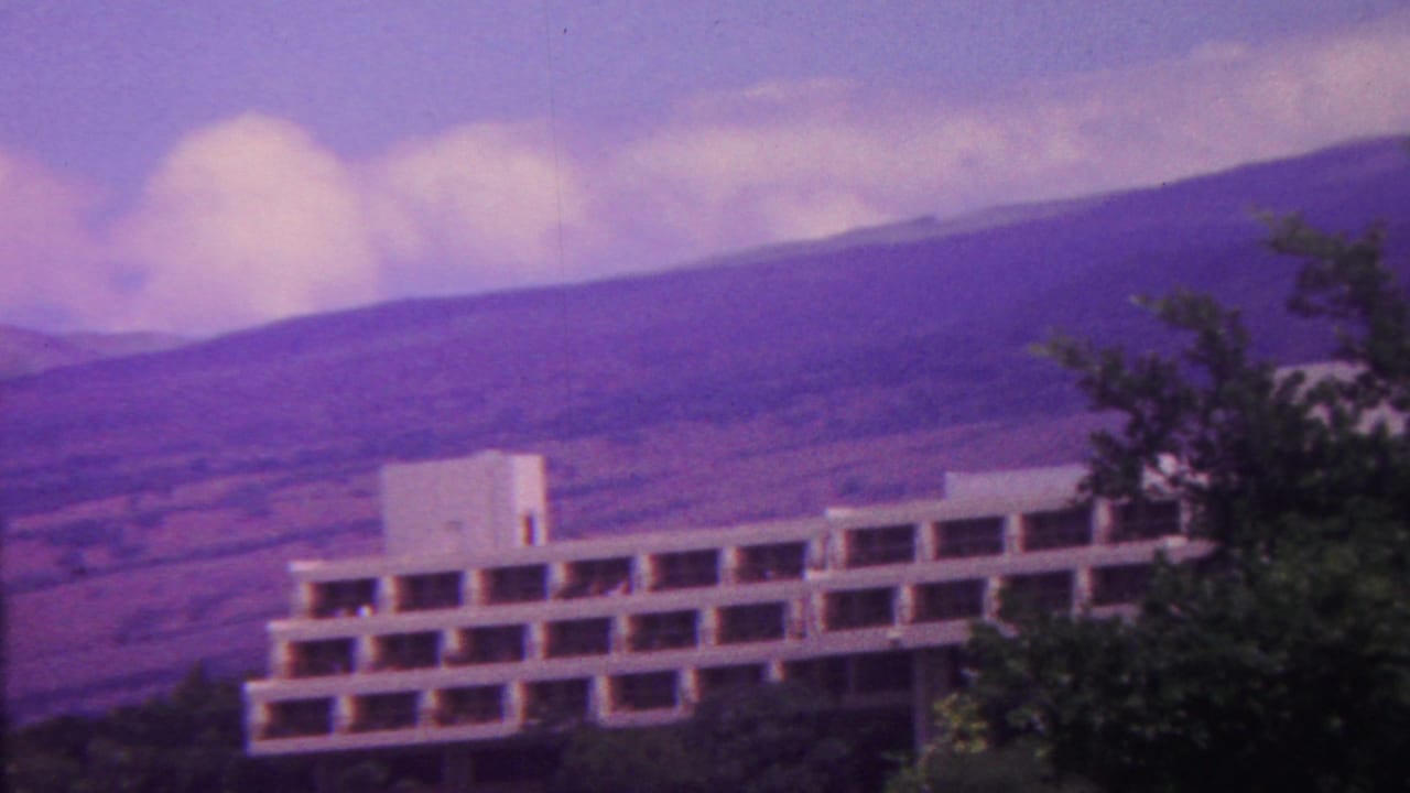 1962 Hawaii 8mm frame showing a white multi-story modernist building partly framed by trees, with volcanic mountain slopes and clouds behind it.