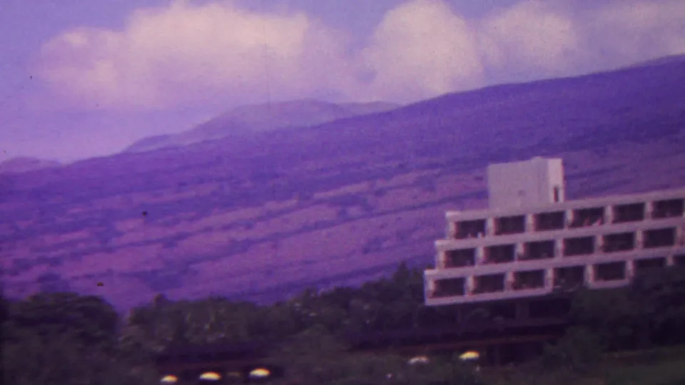 1962 Hawaii 8mm frame with the modernist building shifted to the right edge, backed by layered volcanic hills, trees, and a purple-tinged sky.