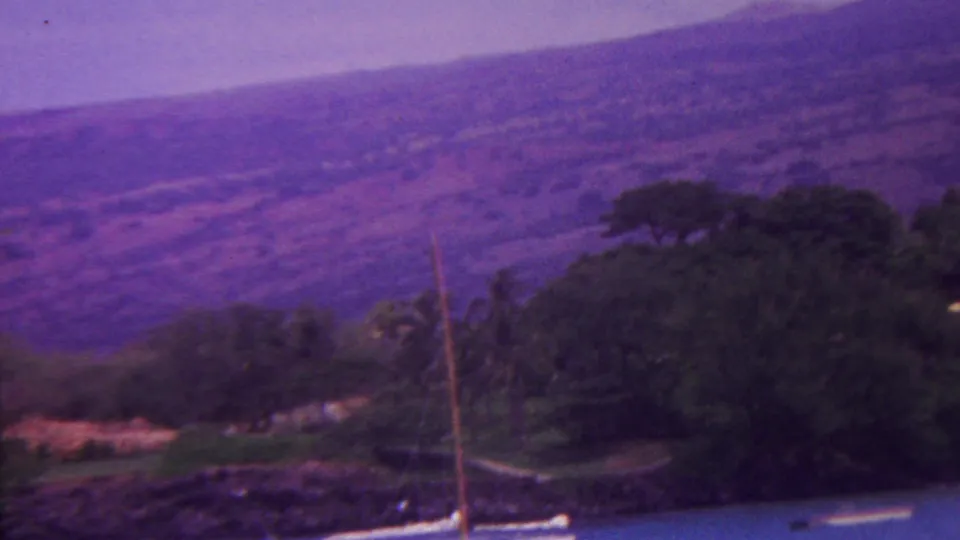 1962 Hawaii 8mm frame showing shoreline water with small boats, dense trees, and purple-toned volcanic slopes in the background.