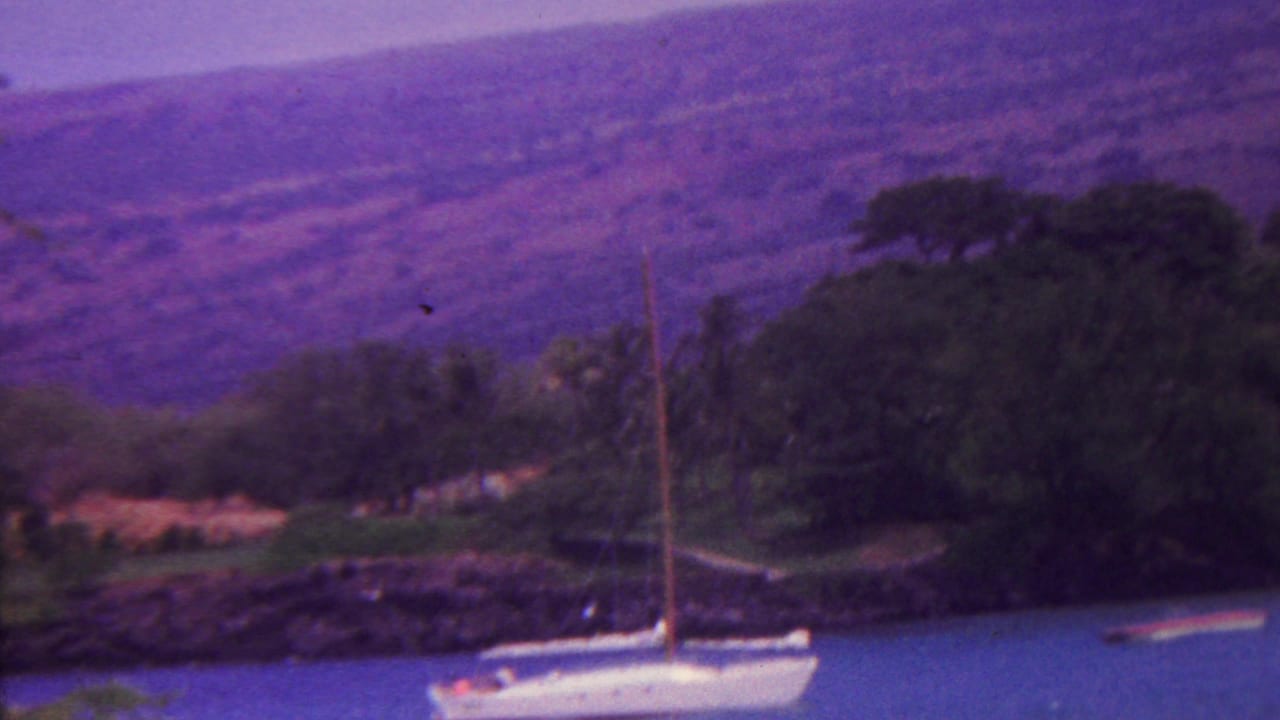 1962 Hawaii 8mm frame showing a white sailboat near a rocky tropical shoreline with dark trees and purple-toned mountains in the background.