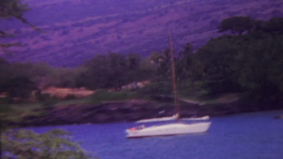 1962 Hawaii coastal view with a white sailboat anchored near dark shoreline rocks, tropical foliage, and hazy purple mountain slopes.