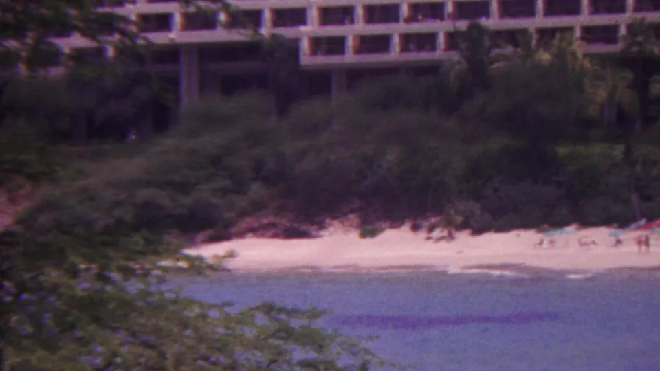 1962 Hawaii shoreline frame showing a pale beach, small distant beachgoers, tropical trees, blue water, and a low resort-like building above the shore.