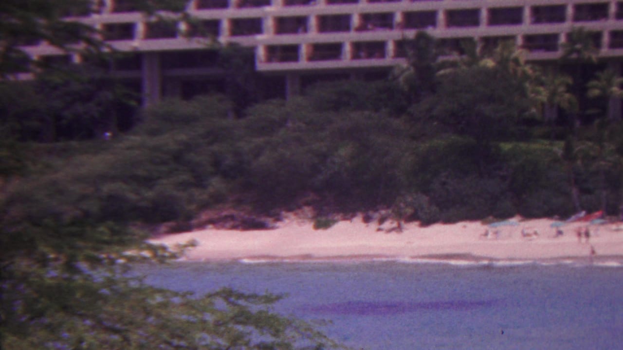 1962 Hawaii home-movie frame showing a large grid-front hotel behind palms and a sandy beach with distant beachgoers beside calm blue water.
