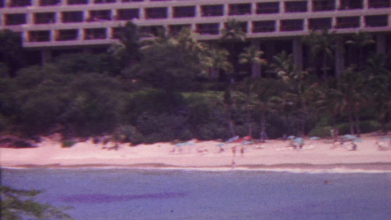 Palm-lined beach below a large hotel with people scattered along the sand and waterline.