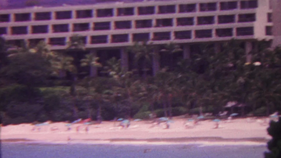 1962 Hawaii beach resort view with the hotel partly cropped at the top, dense palms, small beach umbrellas, and distant people along the shoreline.