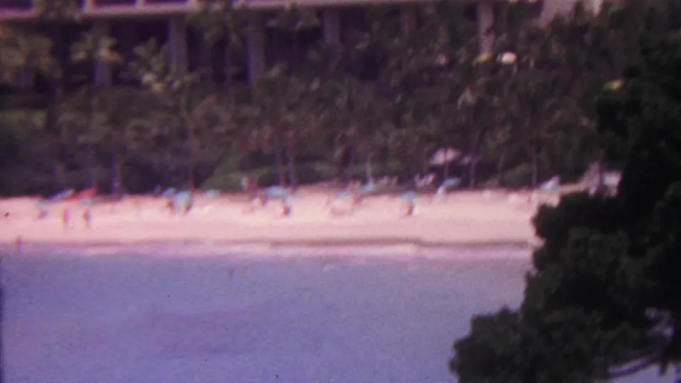 1962 Hawaii home-movie frame showing a palm-backed resort beach with distant beachgoers, blue water, and dark foliage framing the lower right foreground.