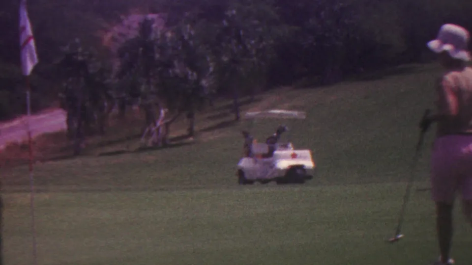 A man in shorts and a wide-brimmed hat stands or walks with a golf club on a green Hawaii golf course near a white golf cart and trees.