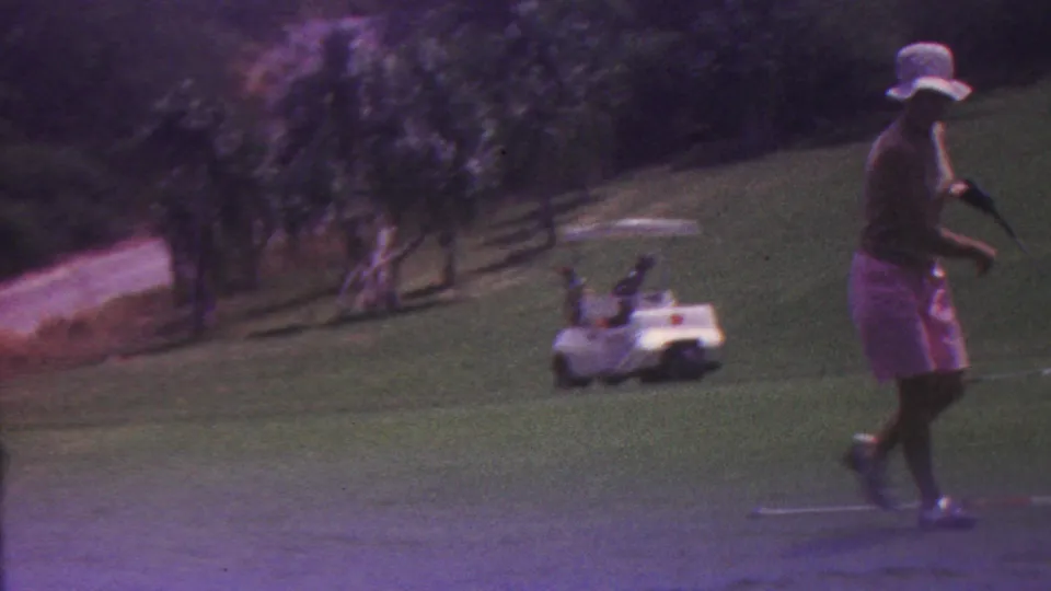 The woman golfer walks across the fairway carrying or holding a club, with a white golf cart, trees, and a course path visible in the background.