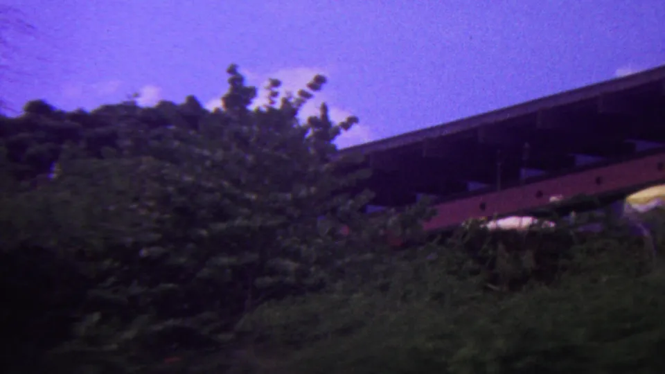1962 Hawaii home-movie frame with tropical greenery in the foreground and a dark elevated structure with a red horizontal element partly visible behind it.