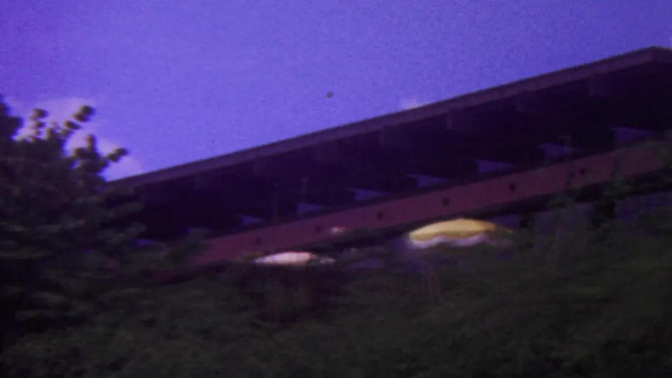 1962 Hawaii 8mm frame showing the camera panning along a dark bridge-like structure above trees, with pale umbrella-like shapes near the greenery.
