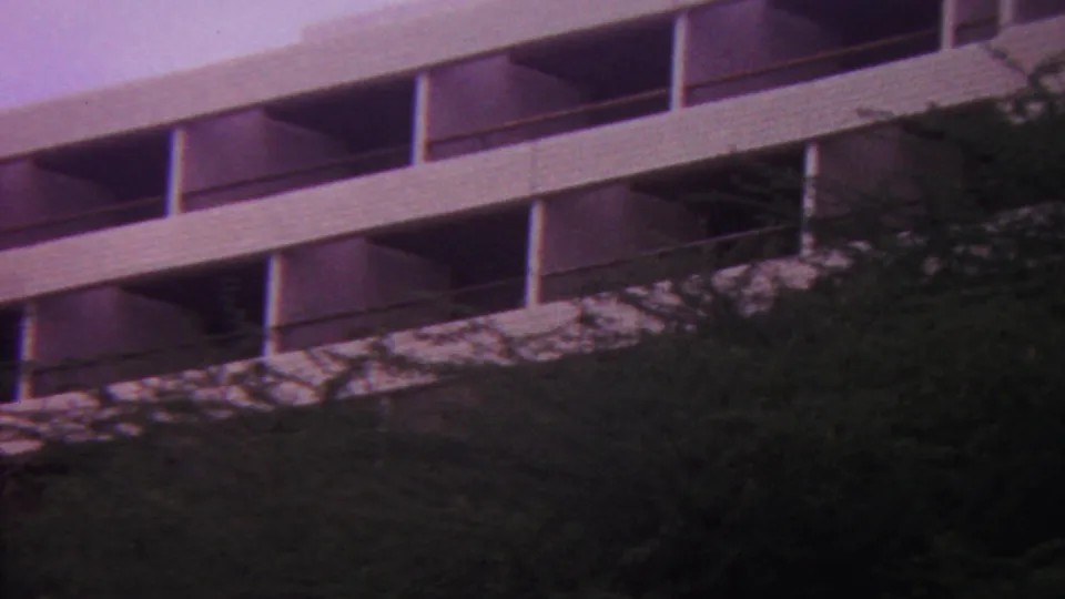 1962 Hawaii home-movie frame revealing a multi-level open concrete parking structure with shaded bays, partly framed by trees and an aged purple film cast.