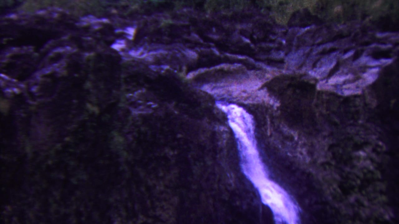 1962 Hawaii 8mm frame showing a narrow white waterfall dropping across dark moss-covered rock, with purple-blue color shift and soft grain.