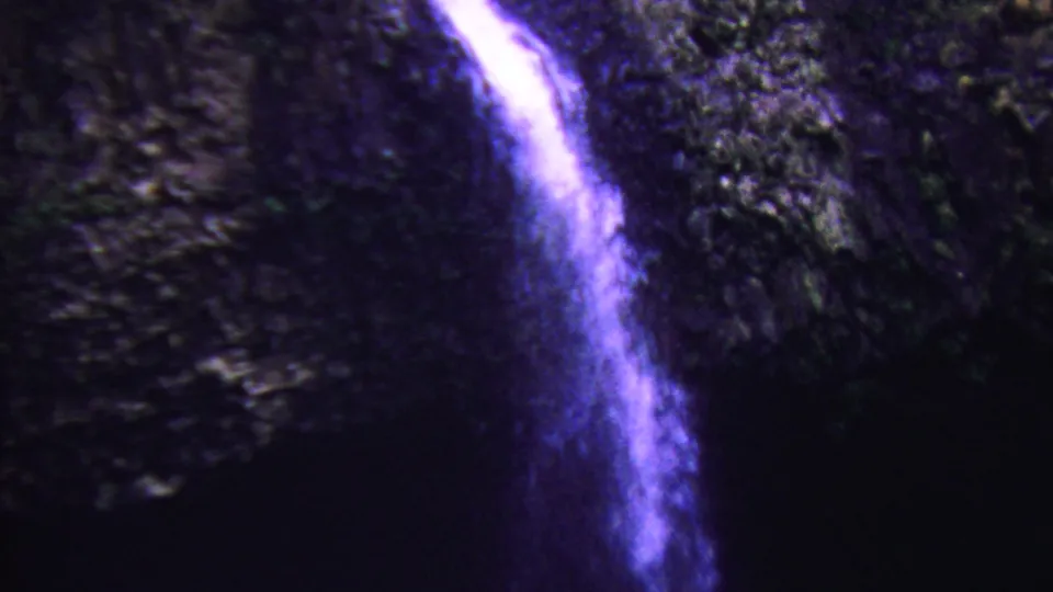 1962 Hawaii 8mm frame showing a tall narrow waterfall descending along a shadowed rocky wall, with purple color drift and soft home-movie texture.