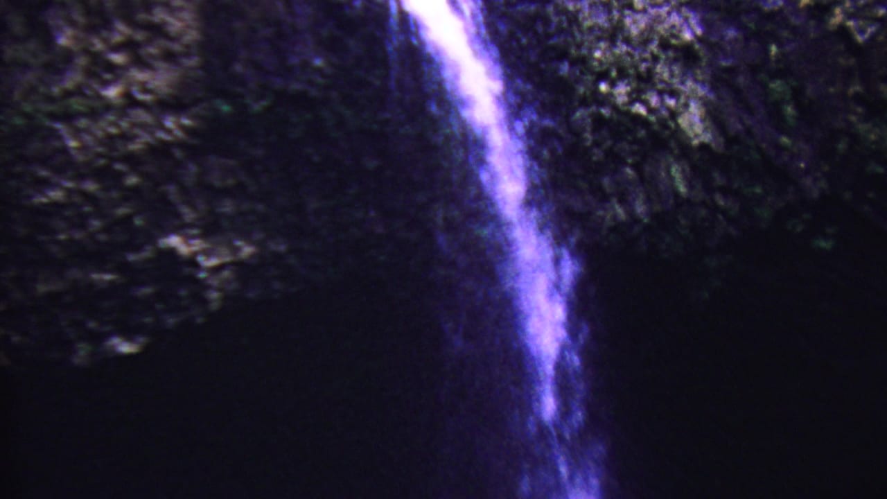 1962 Hawaii 8mm frame showing narrow waterfall streams falling through a dark cave-like rock setting with a strong purple-blue color cast.