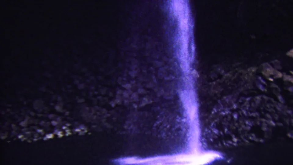 1962 Hawaii 8mm frame showing a narrow waterfall column splashing into a dark pond near rough cave-like rock, tinted by strong purple-blue film color shift.