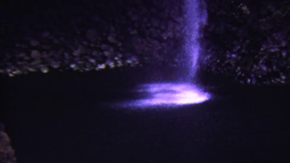 1962 Hawaii home-movie frame showing a purple-lit waterfall with luminous spray below a dark, textured rock formation.