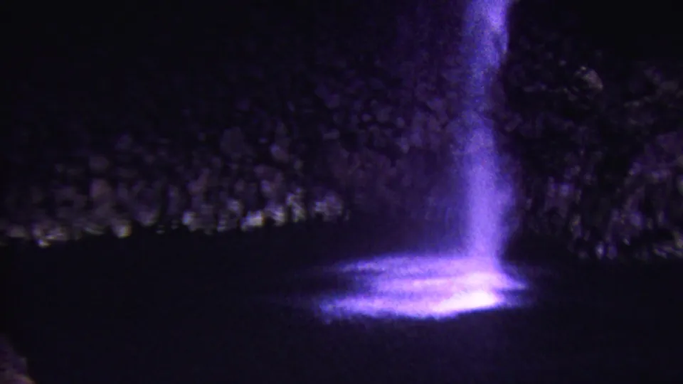 1962 Hawaii 8mm frame of a night waterfall lit in violet, with a bright splash pool and dark rock texture behind it.