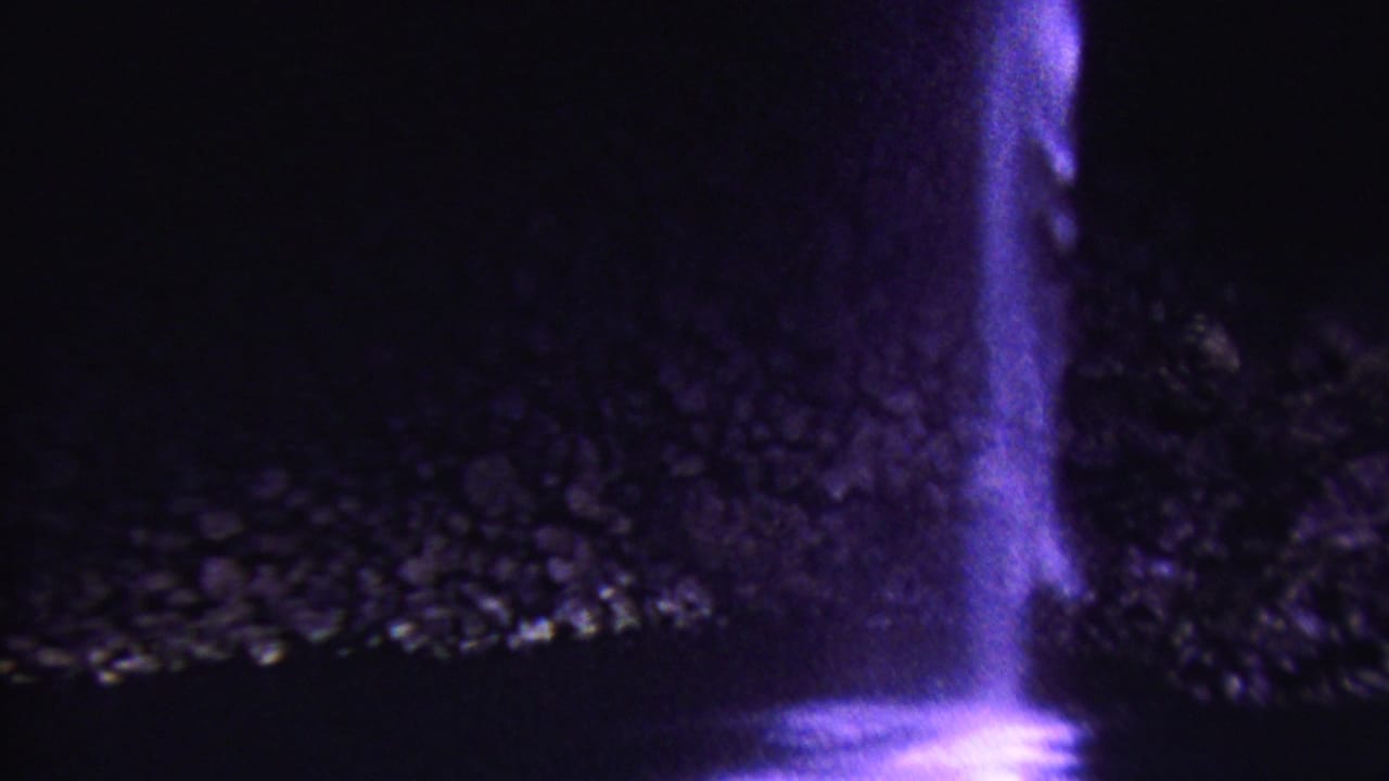 1962 Hawaii 8mm frame showing a narrow waterfall glowing purple against a dark rocky cliff face, with the lower pool mostly obscured by shadow.