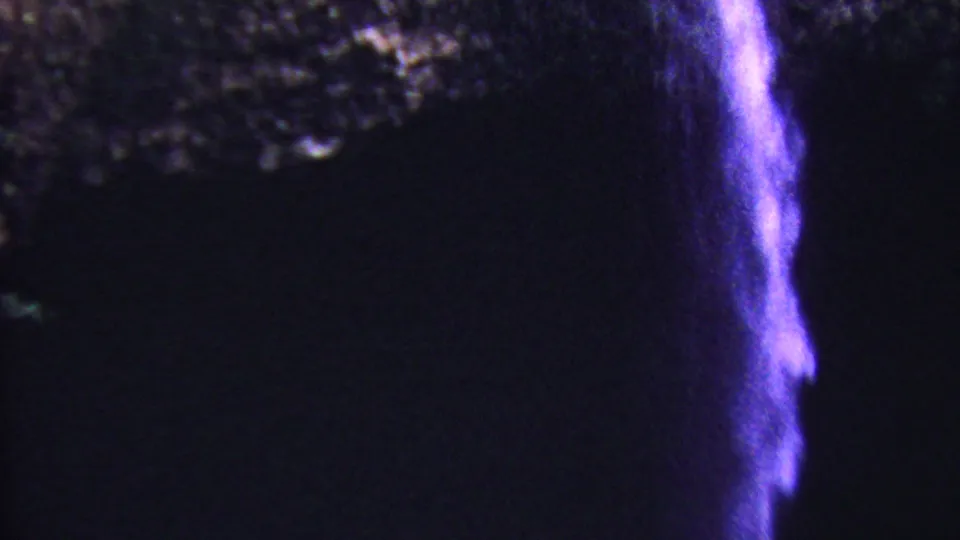 1962 Hawaii 8mm frame showing a purple-lit cascade falling beside a dark basin, with rough rock ledges and low-light film grain visible.
