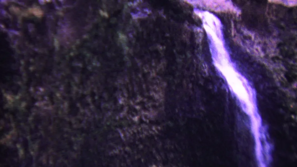1962 Hawaii 8mm frame with a brighter purple-white stream cutting down a rugged rock face, surrounded by dark foliage-like texture and shadow.