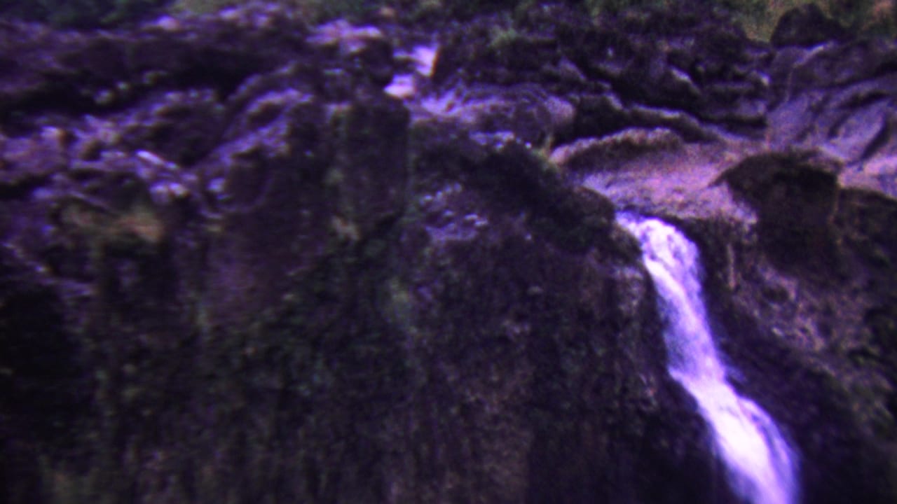 1962 Hawaii 8mm frame showing a narrow white waterfall descending over dark volcanic rock with mossy vegetation and a strong purple color cast.